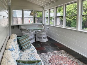 A sunroom with a glass table and wicker chairs, a cushioned bench, and a patterned rug.