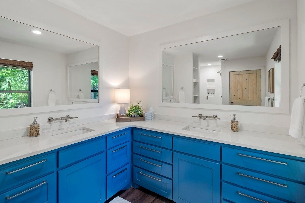 A bathroom with two sinks, blue cabinets, a large mirror, and a window with a view of trees.