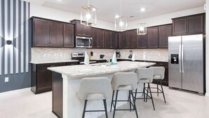 Une cuisine moderne avec des meubles en bois foncé, un réfrigérateur en acier inoxydable, un four encastré et un comptoir en granit. On y trouve trois tabourets de bar et des luminaires suspendus au-dessus de l’îlot central.