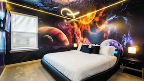 A bedroom with a bed, a nightstand, a lamp, and a wall mural of space.