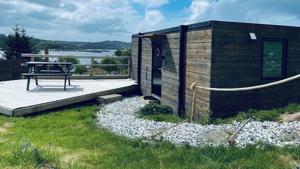Uma cabana moderna com fachada de madeira, um deck com mesa e cadeiras, e vista para o lago e montanhas ao fundo.