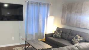Ein Wohnzimmer mit einem grauen Ecksofa, einem flachbildfernseher an der Wand, einem Couchtisch und einem gemusterten Teppich.