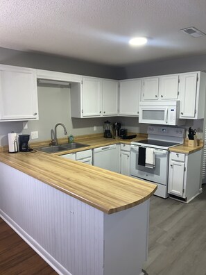 Una cocina con armarios blancos, encimera de madera, un lavamanos, microondas, estufa y lavavajillas.