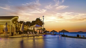A poolside area with lounge chairs and umbrellas, a building with a unique curved facade, and a view of the ocean at sunset.
