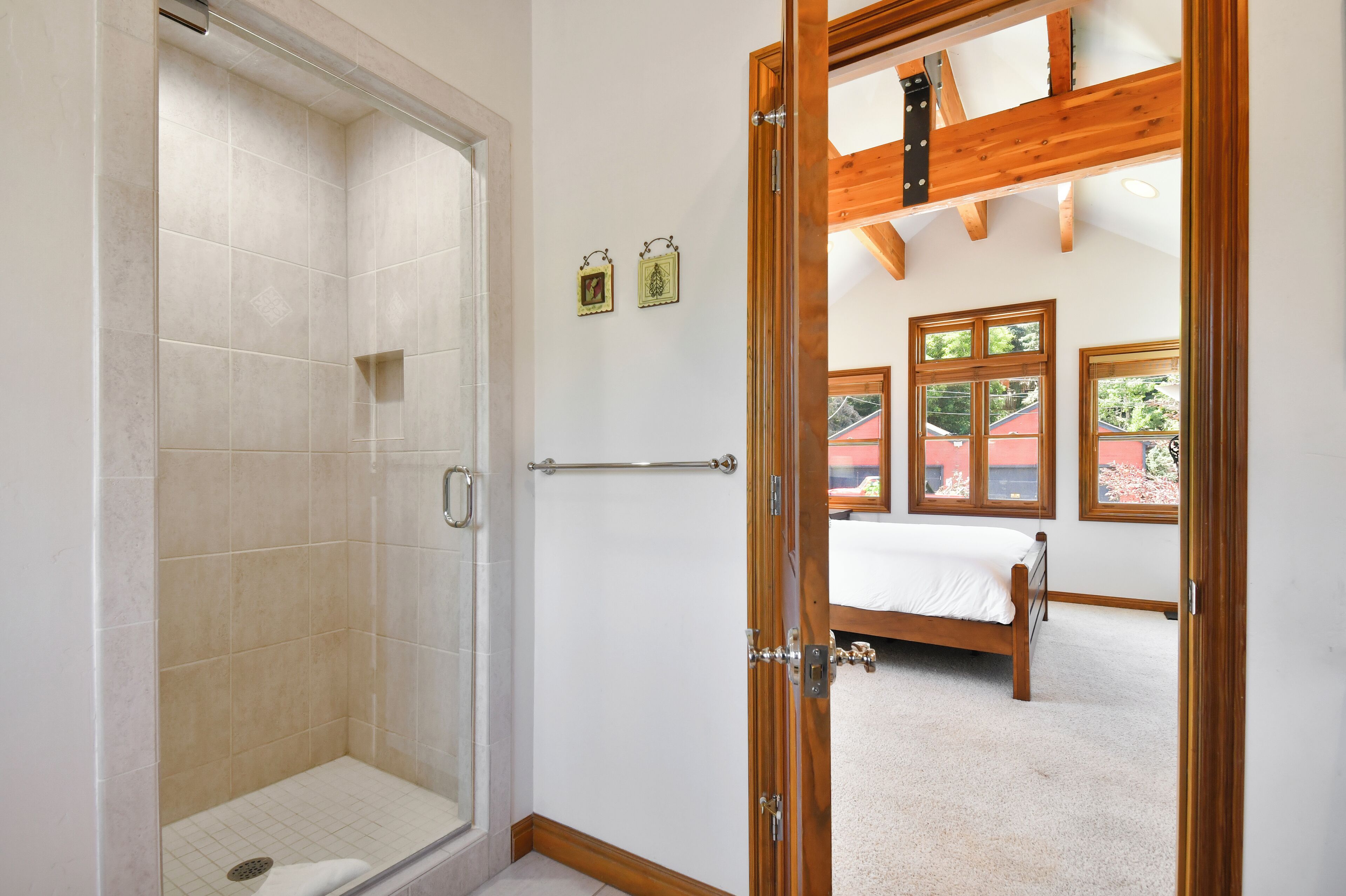 A bathroom with a shower, a toilet, and a bidet. There is a bed with a wooden headboard and a window with a view of a red building outside.
