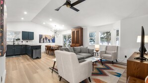 A modern living room with a grey sofa, a coffee table, and a TV stand. There is a dining area with a table and chairs, and a kitchen with dark cabinets and a refrigerator. - Perfectly located home between ski resort and downtown Whitefish- AC for hot days (Whitefish)
