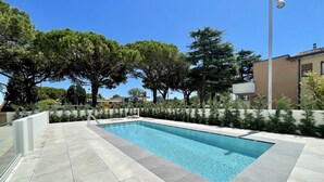 A rectangular swimming pool with a tiled deck, surrounded by trees and a low wall.