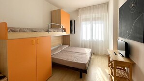 Habitación pequeña con literas, un escritorio y una silla, televisión y un ventanal con cortinas translúcidas.