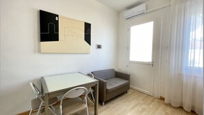 Una habitación con una mesa y sillas blancas, un sofá gris, un ventanal con cortinas blancas y un aire acondicionado instalado en la pared.