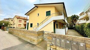 A two-story residential building with a balcony, a stone wall in front, and a gated entrance.