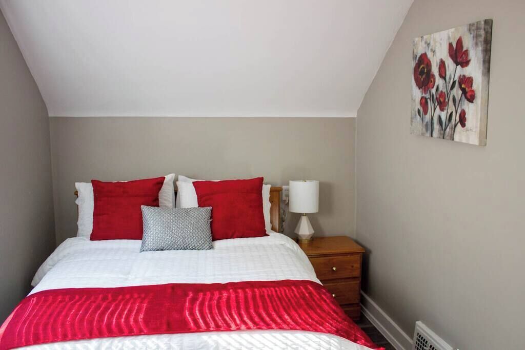 A bedroom with a bed, red and white bedding, a nightstand, a lamp, and a painting on the wall.