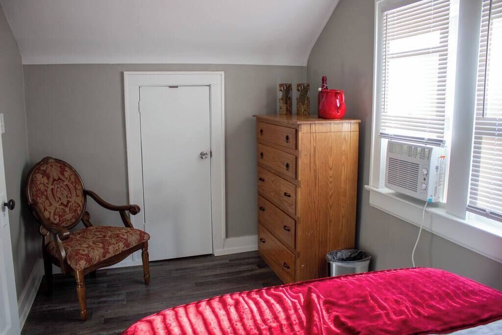 A bedroom with a wooden dresser, a chair, a bed with a red blanket, a door, and a window with blinds.