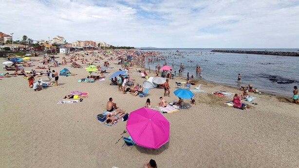 Praia movimentada com pessoas tomando sol e nadando, e edifícios ao fundo.