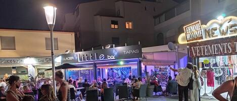 Un espacio para comer al aire libre con mesas y sillas, un bar llamado 'Little bar' y una tienda de ropa llamada 'VETEMENTS'.
