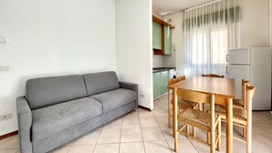 Un espacio habitable compacto con sofá gris, mesa de comedor de madera y sillas, zona de cocina con armarios verdes y un refrigerador blanco.