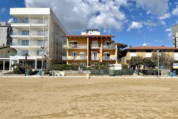 Ein Hotel direkt am Strand mit mehreren Balkonen, einem Sandstrand davor und blauem Himmel.