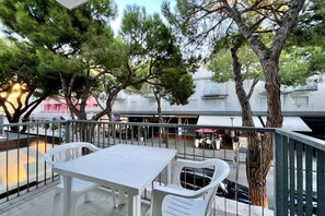 An outdoor patio with a white table and chairs, surrounded by trees and a view of a street with buildings in the background.