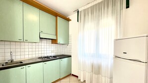 Una cocina con armarios verde claro, un refrigerador blanco y un ventanal con cortinas blancas.