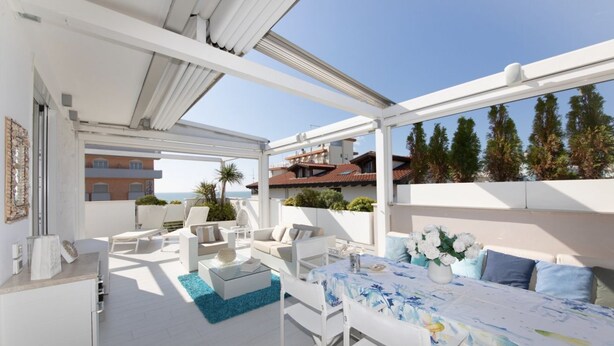 A sunlit outdoor patio with white furniture, a dining table, and a view of the sea and buildings in the background.