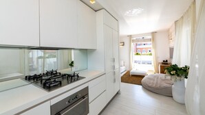 A modern kitchen with white cabinetry, a gas stove, and an oven. There is a large window with curtains, a white sofa, and a plant in the living area.