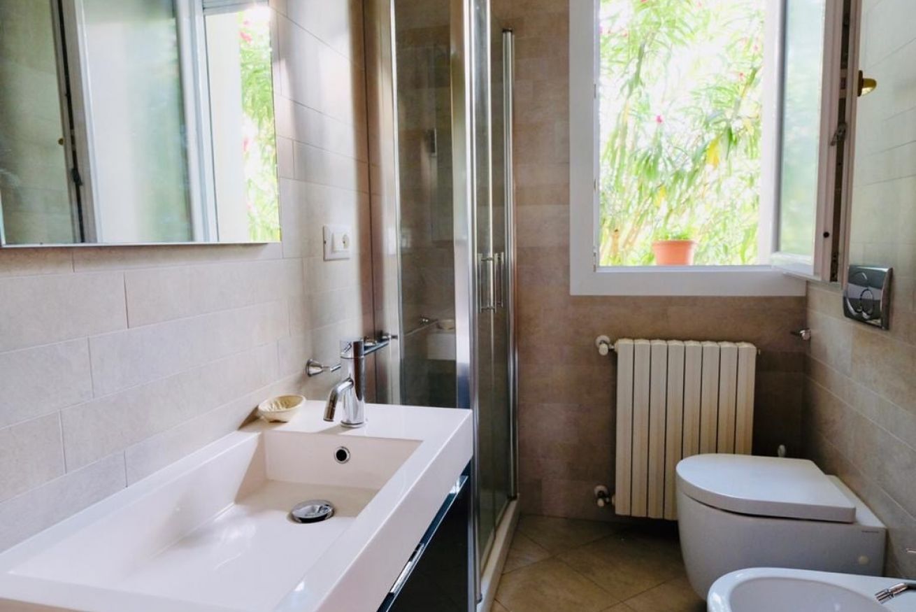 A modern bathroom with a large white bathtub, a glass shower enclosure, a toilet, and a window with a view of greenery.