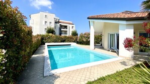 Ein Schwimmbecken mit Fliesenbord, umgeben von Grünpflanzen und einem Gebäude im Hintergrund mit einem ebenfalls gefliesten Dach.