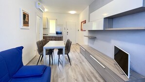 A modern living room with a blue sofa, a dining table with chairs, a TV, and a kitchen in the background.