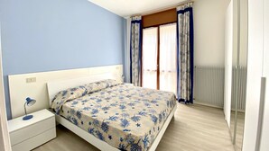 A bedroom with a bed, nightstand, and a window with curtains. - Appartamento Villa Santin 4-Carraro Immobiliare Jesolo (Lido di Jesolo)