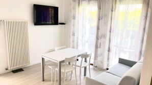 A modern living room with a white sofa, a dining table with chairs, a radiator, and a TV mounted on the wall. - Appartamento Villa Santin 4-Carraro Immobiliare Jesolo (Lido di Jesolo)