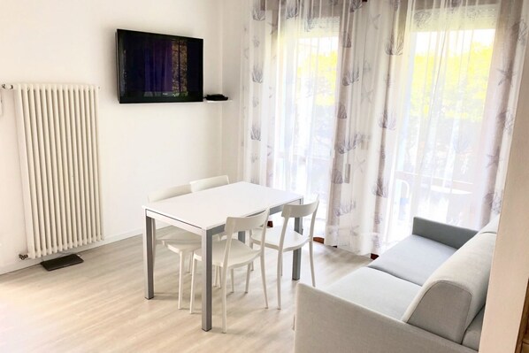 A modern living room with a white sofa, a dining table with chairs, a radiator, and a TV mounted on the wall.
