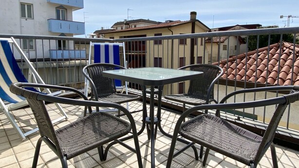 A balcony with wicker chairs and a glass table, overlooking a residential area with buildings and a clear sky. - Appartamento Villa Santin 4-Carraro Immobiliare Jesolo (Lido di Jesolo)