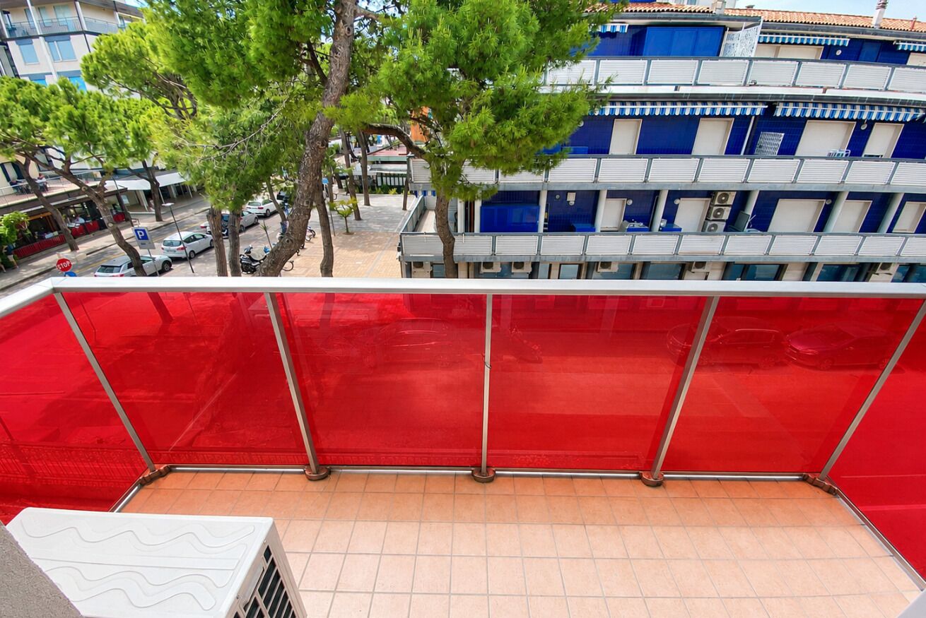 En balkon med rødt sikkerhedsnet, en hvid aircondition og udsigt til en blå og hvid bygning med flere balkoner.