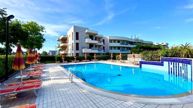 Ein Schwimmbecken mit Liegestühlen und Sonnenschirmen, umgeben von einer gefliesten Terrasse und modernen Gebäuden im Hintergrund.