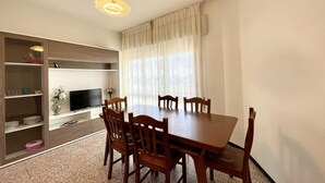 Un área de comedor con una mesa y sillas de madera, un televisor en un estante y una ventana con cortinas traslúcidas.