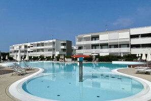 A swimming pool with lounge chairs and umbrellas, surrounded by modern buildings with balconies. - Appartamento Laguna - Carraro Immobiliare Jesolo (Jesolo)