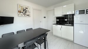 A modern kitchen with white cabinets, a black countertop, and a white refrigerator. There is a dining table with black chairs and a flat-screen TV mounted on the wall. - DB1 Appartamento 8 posti letto Carraro Immobiliare Jesolo (Lido di Jesolo)