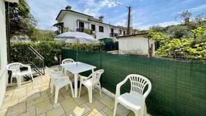 Um pátio com cadeiras e mesa de plástico branco, cerca verde e um edifício branco ao fundo.