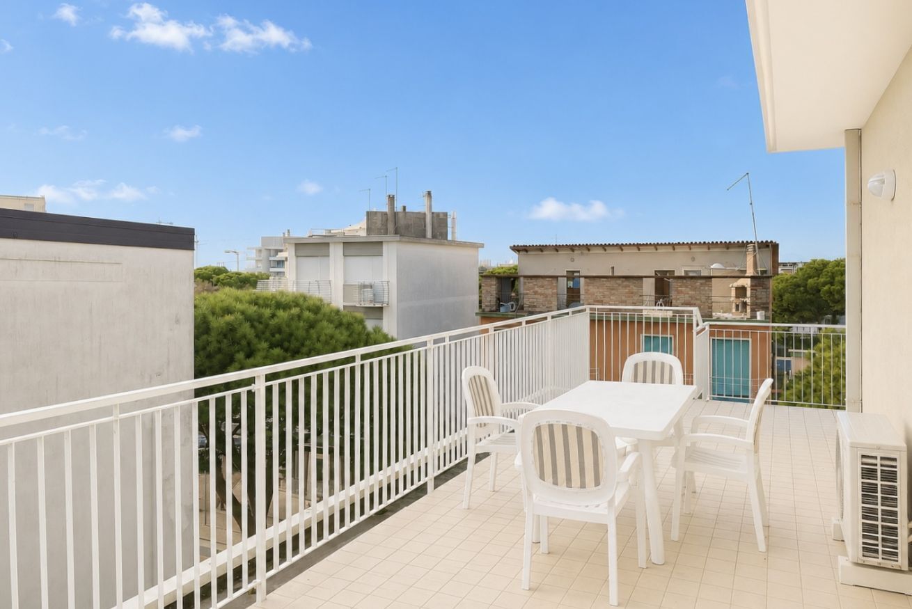 Uma varanda com uma mesa e cadeiras brancas, vista para edifícios vizinhos e céu claro.
