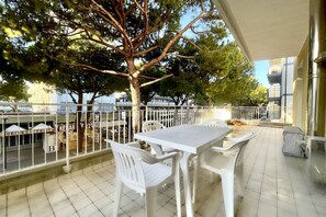 Un balcón con una mesa y sillas blancas, un árbol y vistas de la calle con carteles que indican 'DRINKS & FRUITS' y 'KEBAB'.