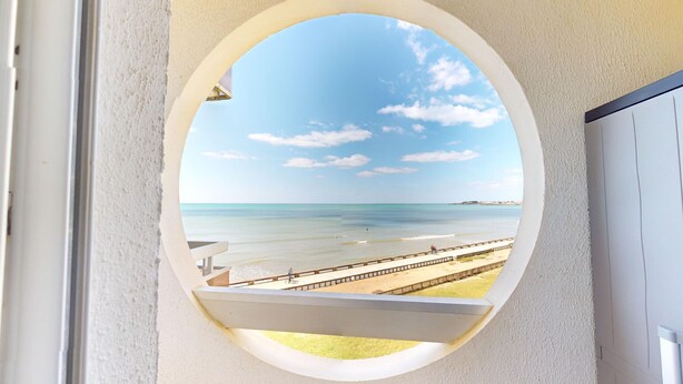A view through a circular window of a beach with a wooden walkway and a distant shoreline. - Panoramic View face Mer (Saint-Gilles-Croix-de-Vie)