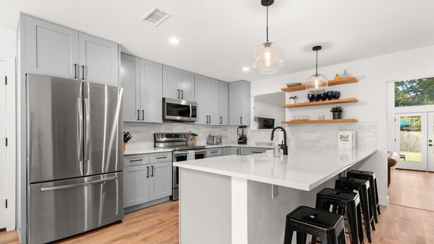 A modern kitchen with grey cabinets, stainless steel appliances, a central island with stools, and wooden shelves above.