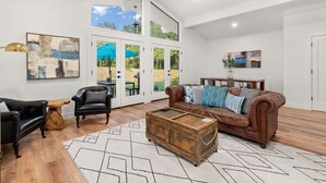 A spacious living room with a brown leather sofa, black armchairs, and a wooden coffee table. Large windows provide natural light and a view of the outdoors.