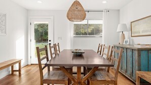 A dining area with a wooden table and chairs, a woven pendant light, and a view through the window.