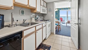 A compact kitchen with a dishwasher, sink, and stove. A dining area with a table and chairs is visible through an open door leading to a balcony with a view of the sea. - Tropical oceanfront resort with shops, restaurants, pools, lazy river-paradise! Sleeps 4. (North Myrtle Beach)