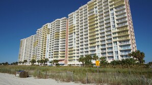 A high-rise hotel with multiple balconies, a sandy beach in the foreground, and a 'Surfing Area' sign. - Tropical oceanfront resort with shops, restaurants, pools, lazy river-paradise! Sleeps 4. (North Myrtle Beach)