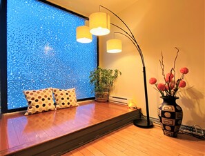 Una habitación con piso de madera, un sofá con cojines de lunares, un ventanal con un muro de mosaico, una lámpara de pie, una planta en maceta y un florero con flores.