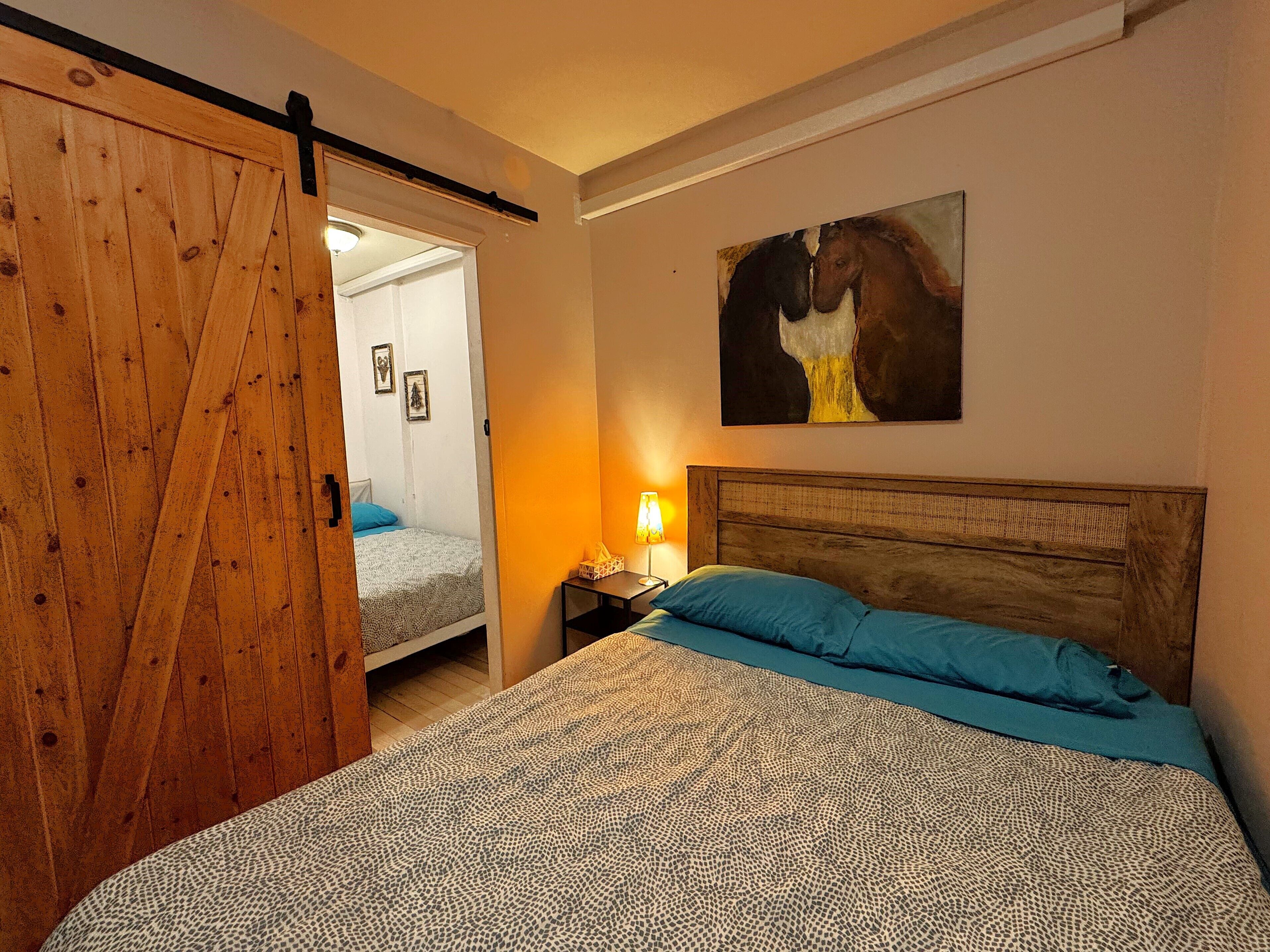 A bedroom with a bed, a nightstand, a lamp, a wooden door, and a painting on the wall.