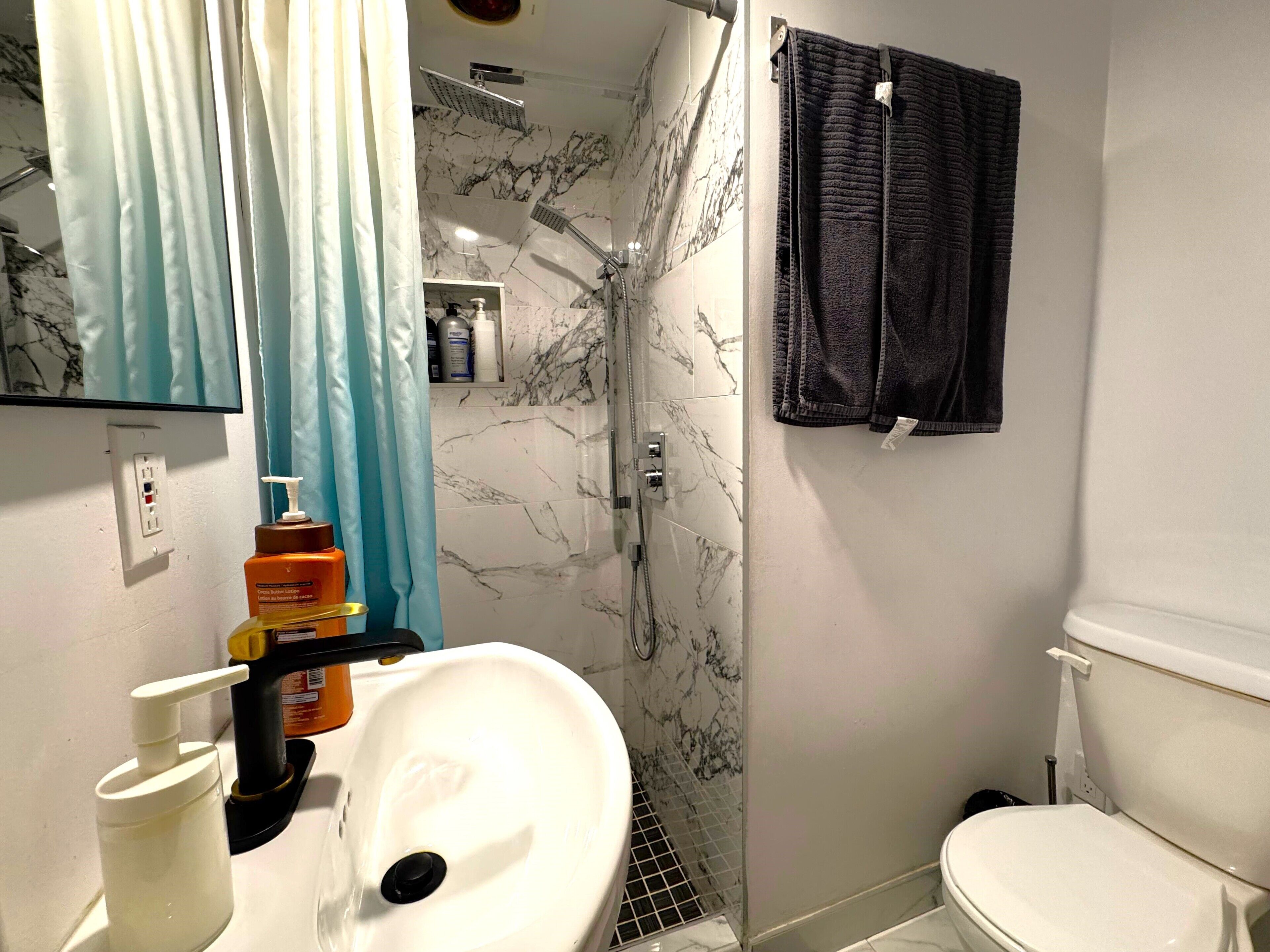 Une salle de bain avec un lavabo blanc, un miroir, une douche avec un mur en marbre et des toilettes.