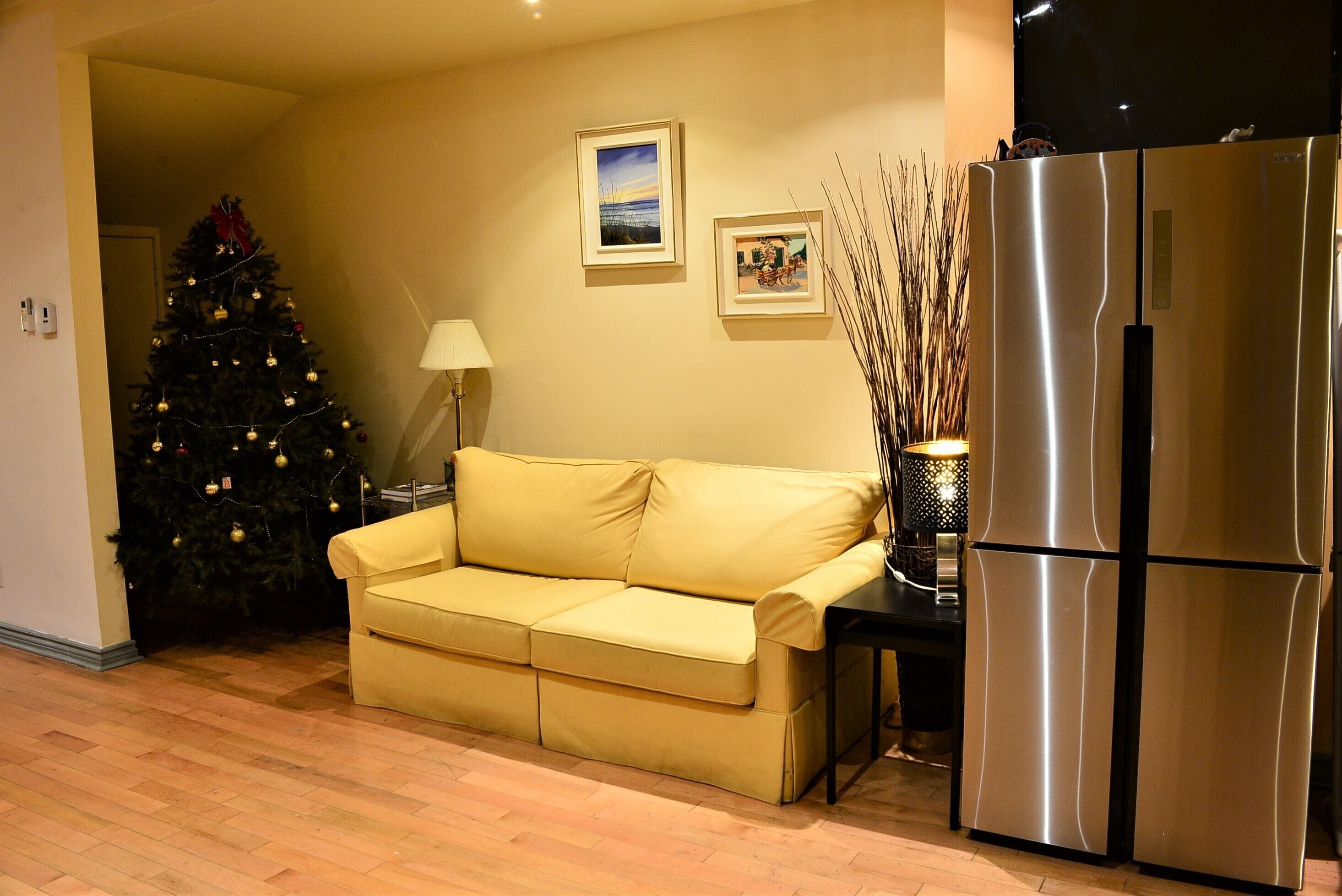 Una sala de estar con un sofá color beige, un árbol de Navidad, un refrigerador y dos cuadros en la pared.