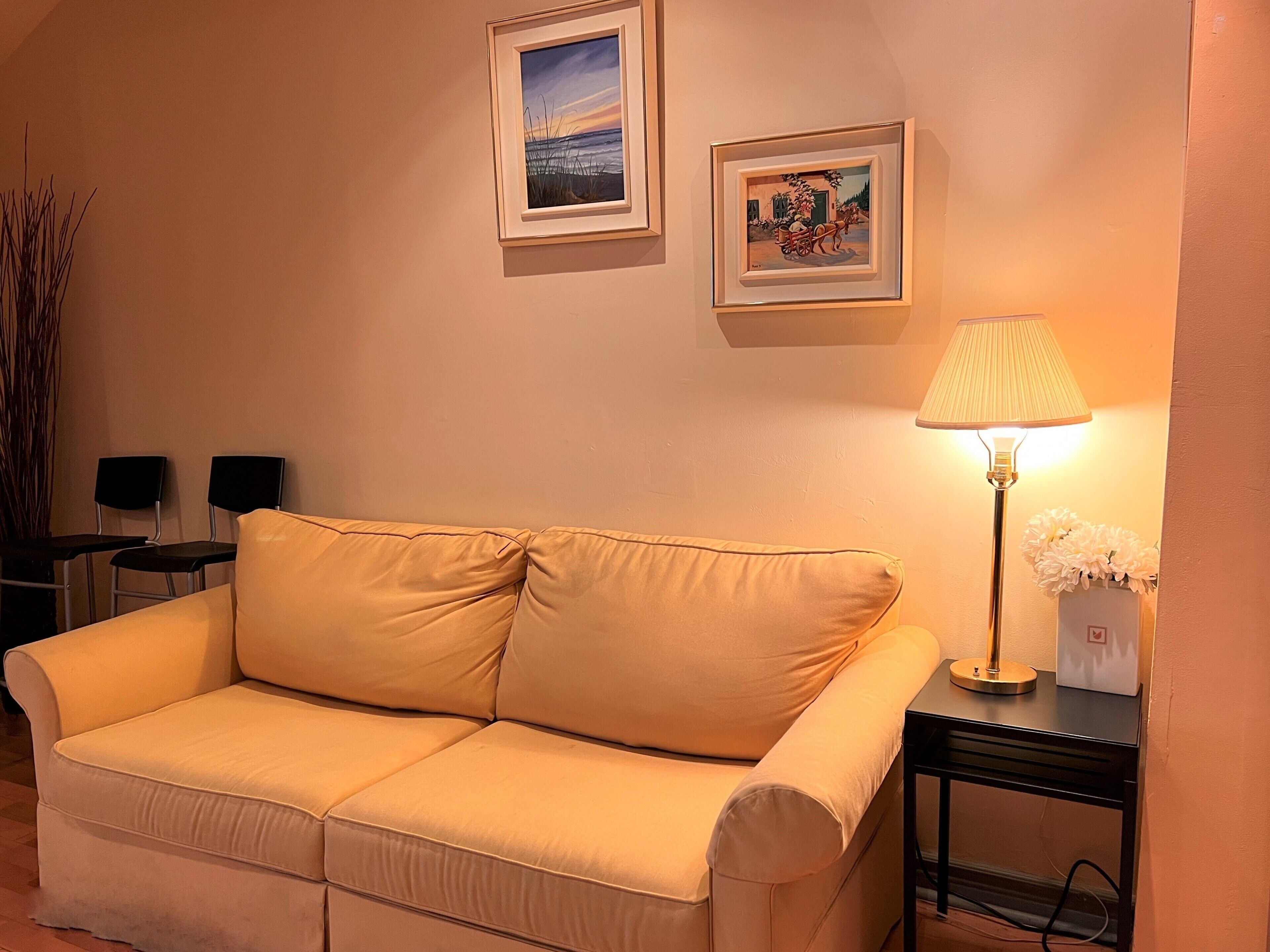 A living room with a beige sofa, two framed artworks on the wall, a lamp, and a vase with flowers on a side table.